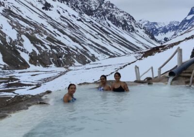 Tres turistas disfrutando de las aguas termales de las Termas de Colina con el paisaje nevado del Cajón del Maipo al fondo.